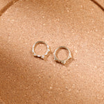 A pair of silver-toned hoop earrings with small clear stones set along the front, displayed on a textured brown surface.