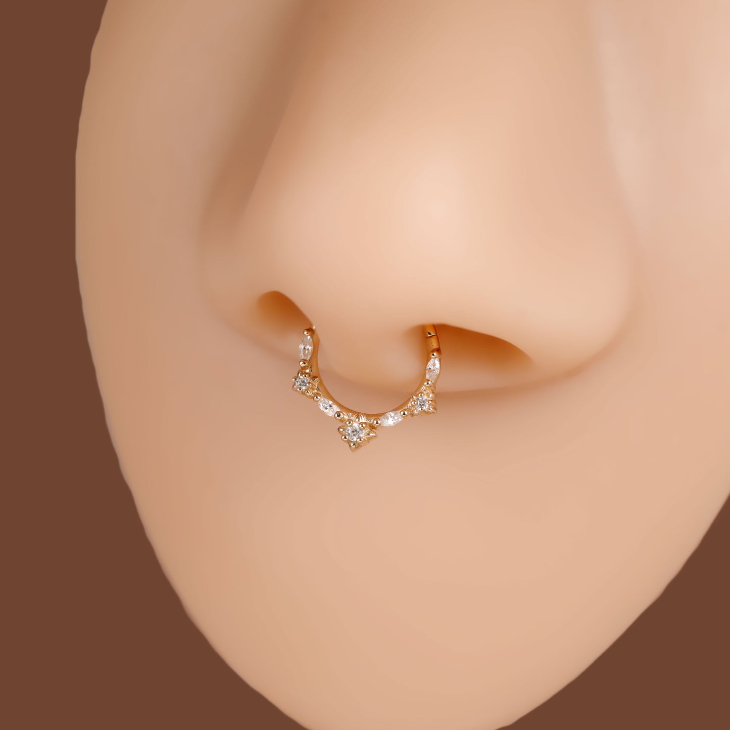 A close-up of a gold crescent nose hoop stud adorned with small clear crystals, shown on a mannequin nose against a solid brown background.