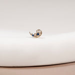 A small gold crescent moon earring with two blue gemstones, displayed on a white curved surface.