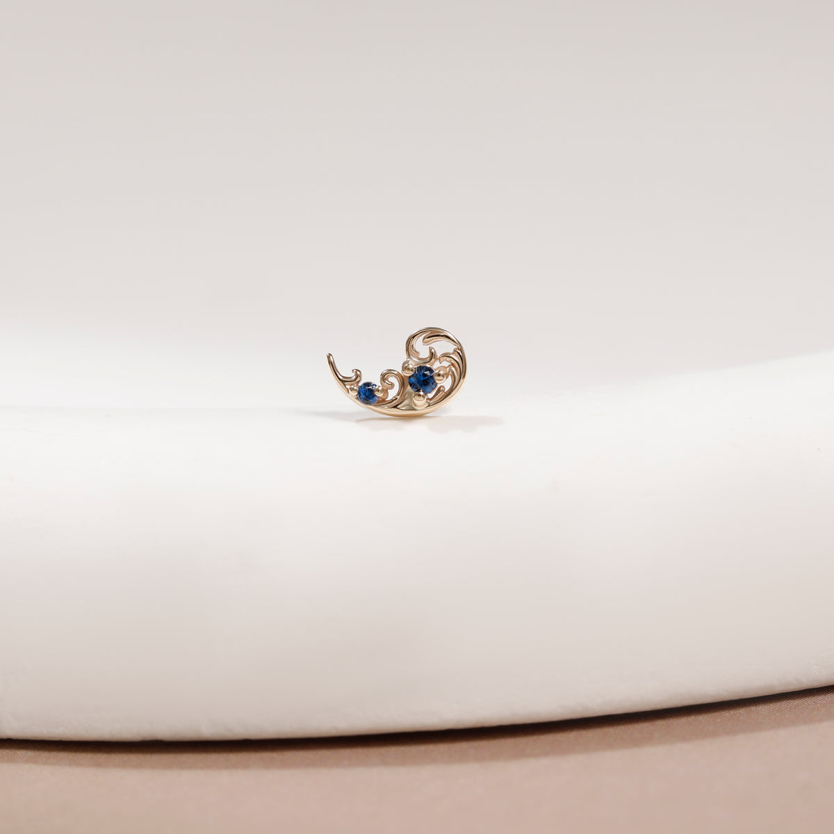A small gold crescent moon earring with two blue gemstones, displayed on a white curved surface.