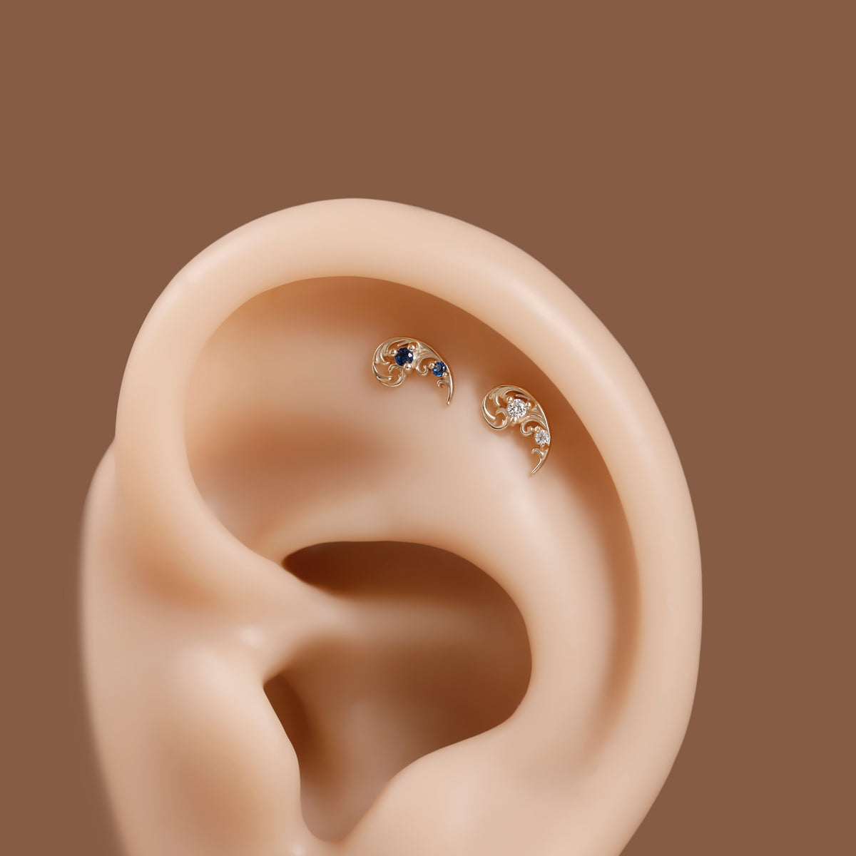 Two small curved ear piercings with gold-toned metal and dark blue gemstones, displayed on a beige ear model against a solid brown background.