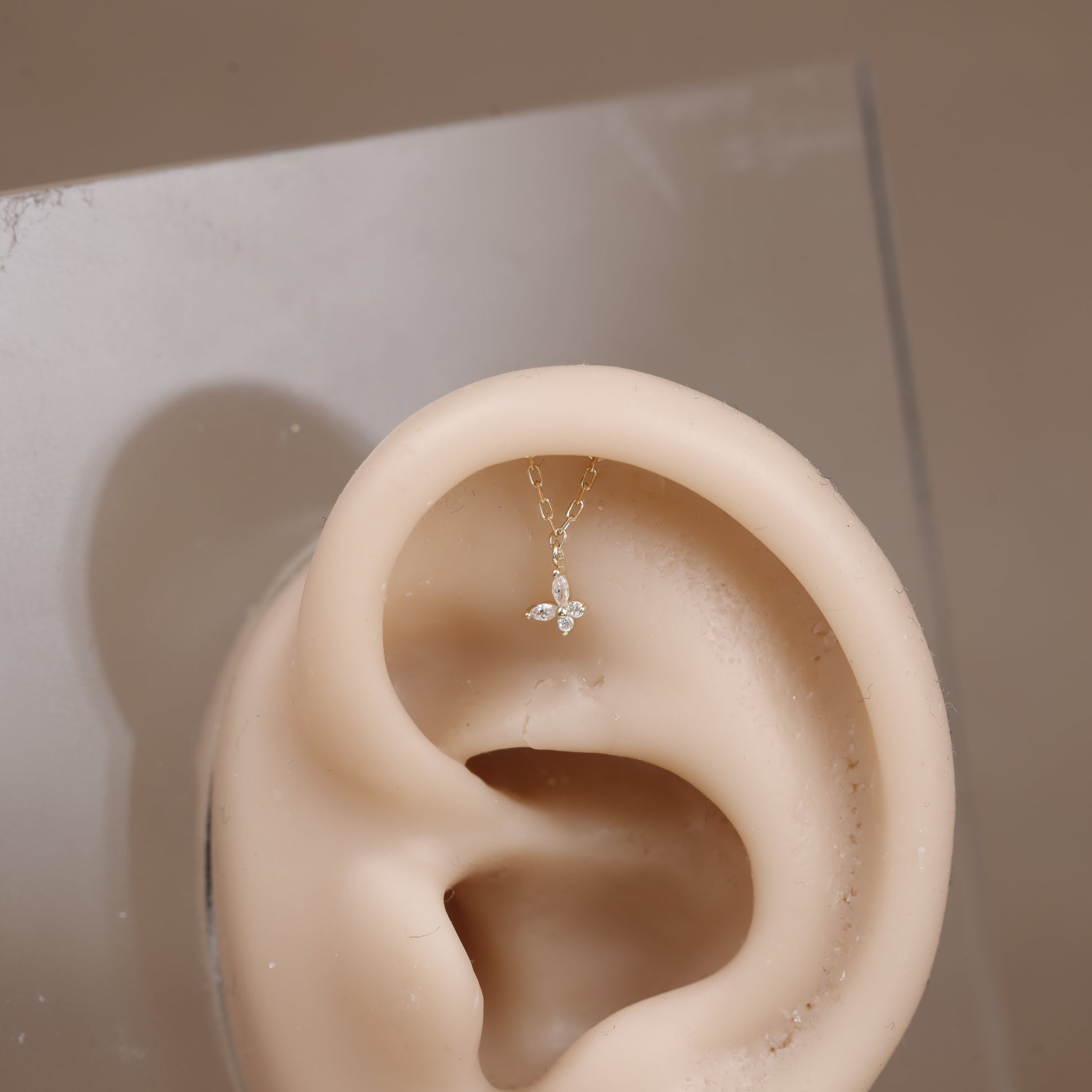 A delicate gold ear cuff necklace with a small butterfly charm set with clear stones, displayed on a beige silicone ear model.