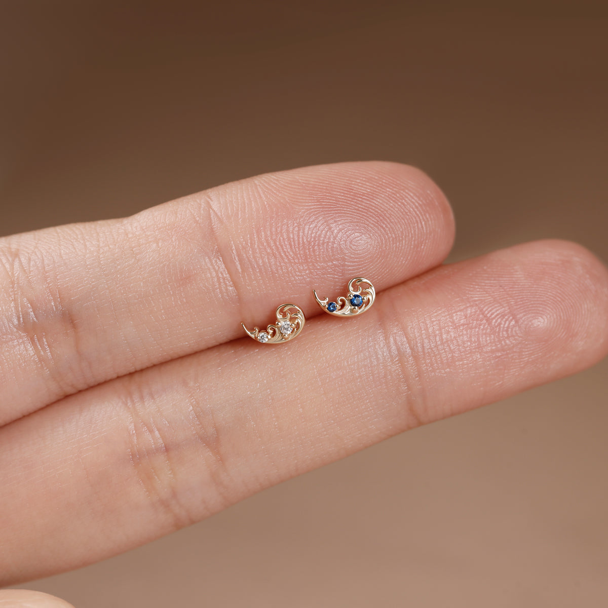 A close-up of a person's fingers displaying a pair of small gold crescent moon stud earrings with tiny blue and clear stones.