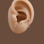 A pair of gold ear cuff piercings with dangling clear crystal teardrop charms, displayed on a beige silicone ear model against a solid brown background.