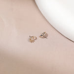 A pair of delicate gold crescent moon stud earrings with tiny clear stones, displayed on a soft beige satin surface next to a white ceramic dish.