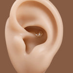 A close-up of a human ear wearing a small gold helix hoop earring with a clear gemstone, set against a solid brown background.