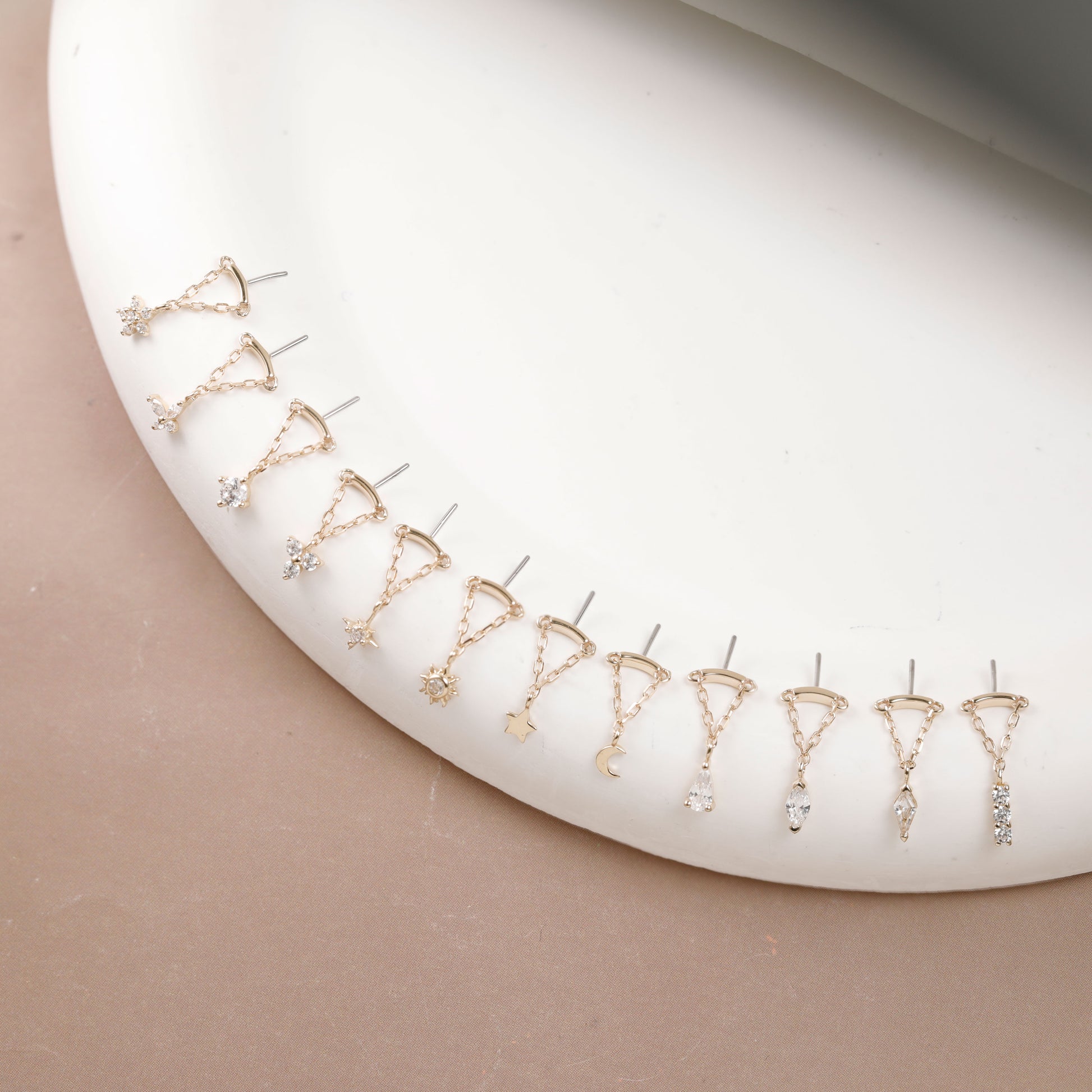 A set of small gold-toned and silver-toned dangle earrings arranged in a curved row on a white oval display stand against a warm beige background.
