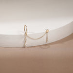 A delicate gold huggie hoop earring with a short chain connecting to two small dangling charms, displayed on a white curved jewelry stand against a warm beige background.