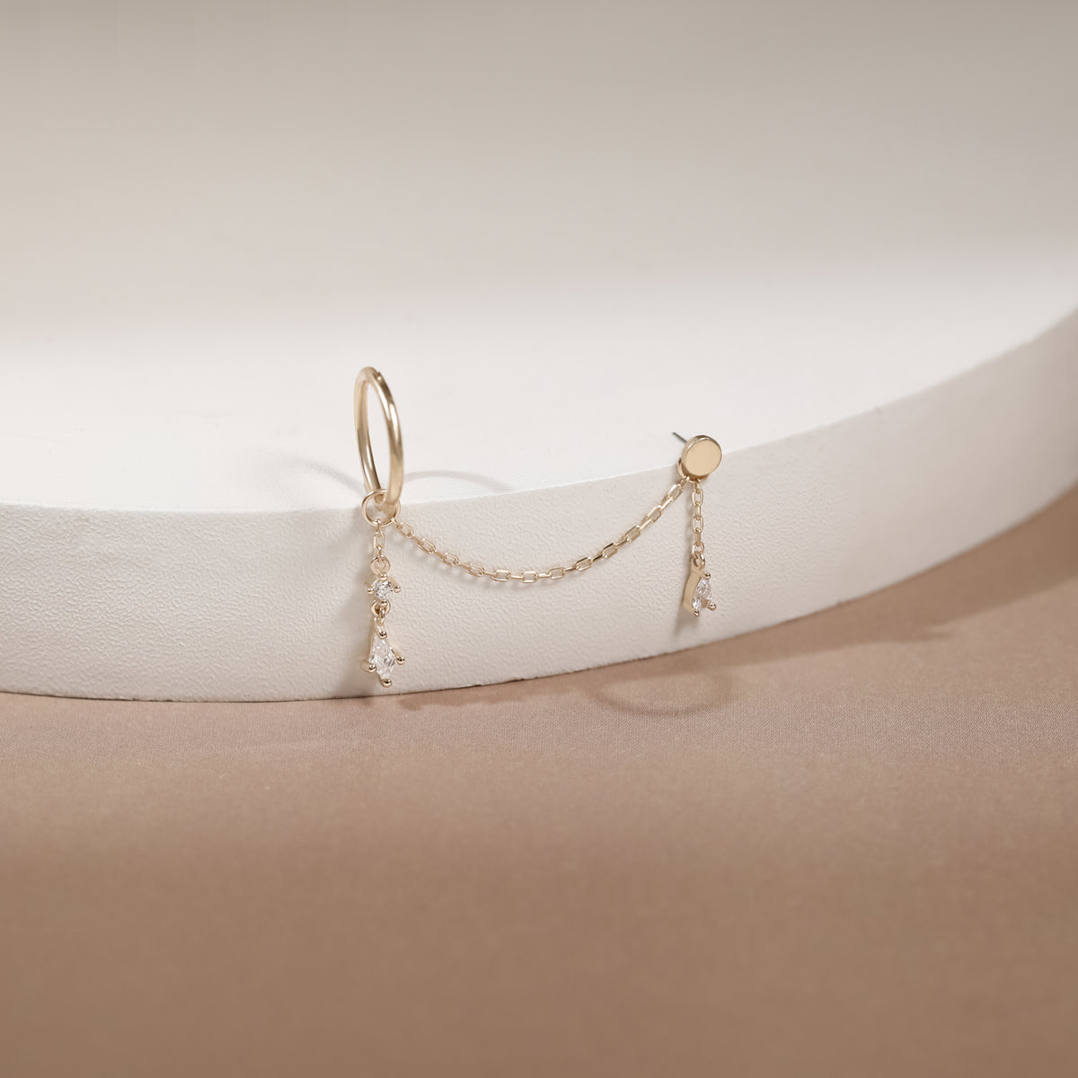 A delicate gold huggie hoop earring with a short chain connecting to two small dangling charms, displayed on a white curved jewelry stand against a warm beige background.