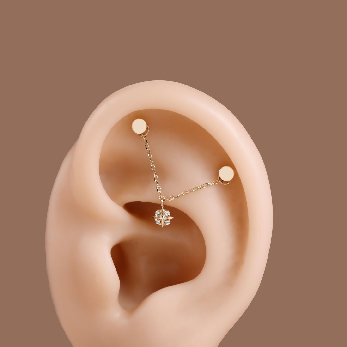 A close-up of a human ear wearing a delicate gold ear cuff with two small gold studs connected by a fine chain, with a small clear gemstone at the center, set against a solid brown background.