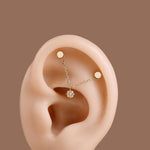 A close-up of a human ear wearing a delicate gold ear cuff with two small gold studs connected by a fine chain, with a small clear gemstone at the center, set against a solid brown background.