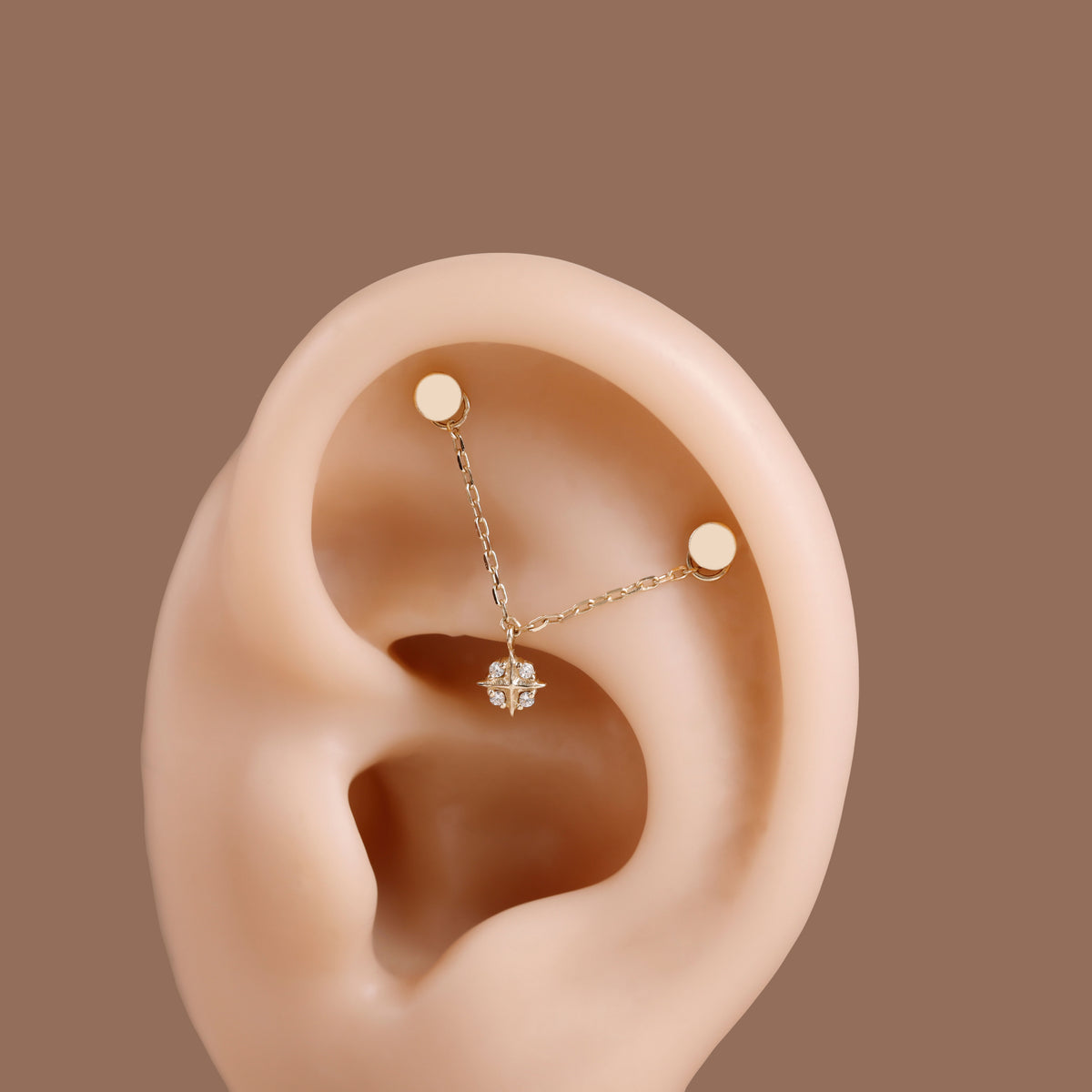 A close-up of a human ear wearing a delicate gold ear cuff with two small gold studs connected by a fine chain, with a small clear gemstone at the center, set against a solid brown background.