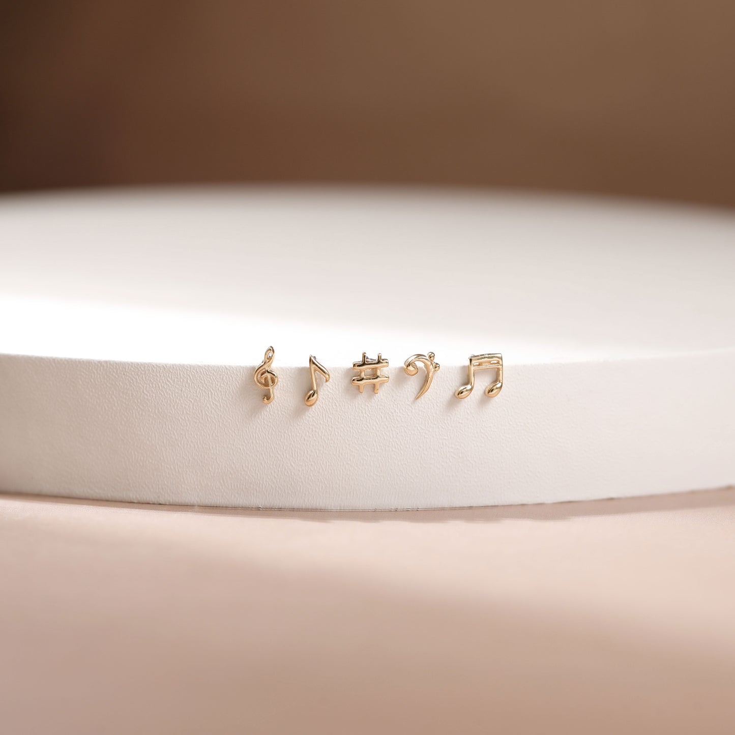 Four small gold stud earrings shaped like musical notes, displayed on a white cylindrical pedestal against a soft beige background.