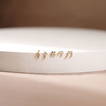 Four small gold stud earrings shaped like musical notes, displayed on a white cylindrical pedestal against a soft beige background.