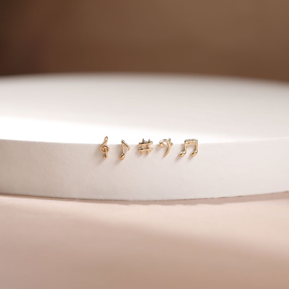 Four small gold stud earrings shaped like musical notes, displayed on a white cylindrical pedestal against a soft beige background.