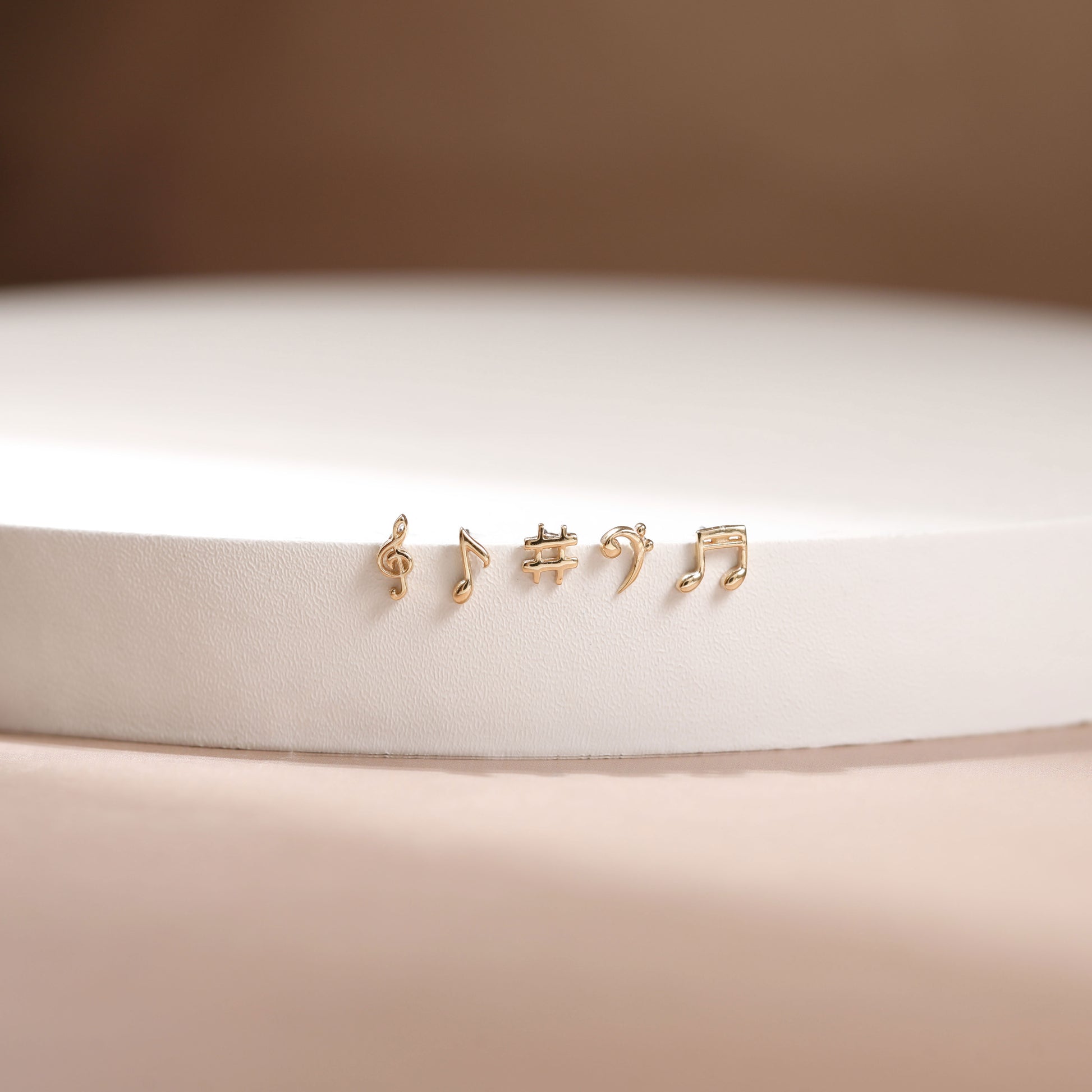 Four small gold stud earrings shaped like musical notes, displayed on a white cylindrical pedestal against a soft beige background.
