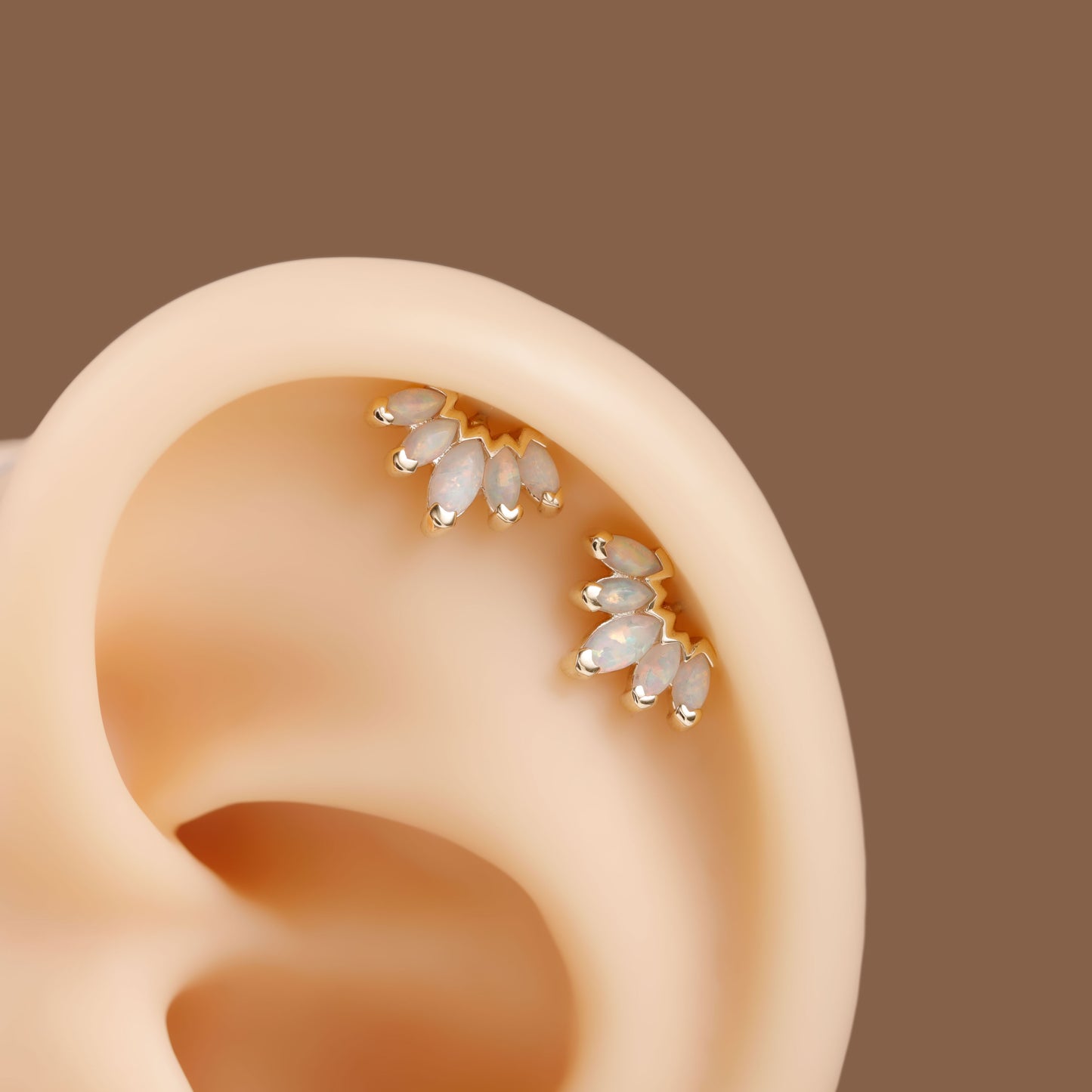 A close-up of a human ear wearing two small gold ear cuff studs with white teardrop-shaped stones, set against a solid brown background.
