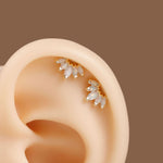 A close-up of a human ear wearing two small gold ear cuff studs with white teardrop-shaped stones, set against a solid brown background.