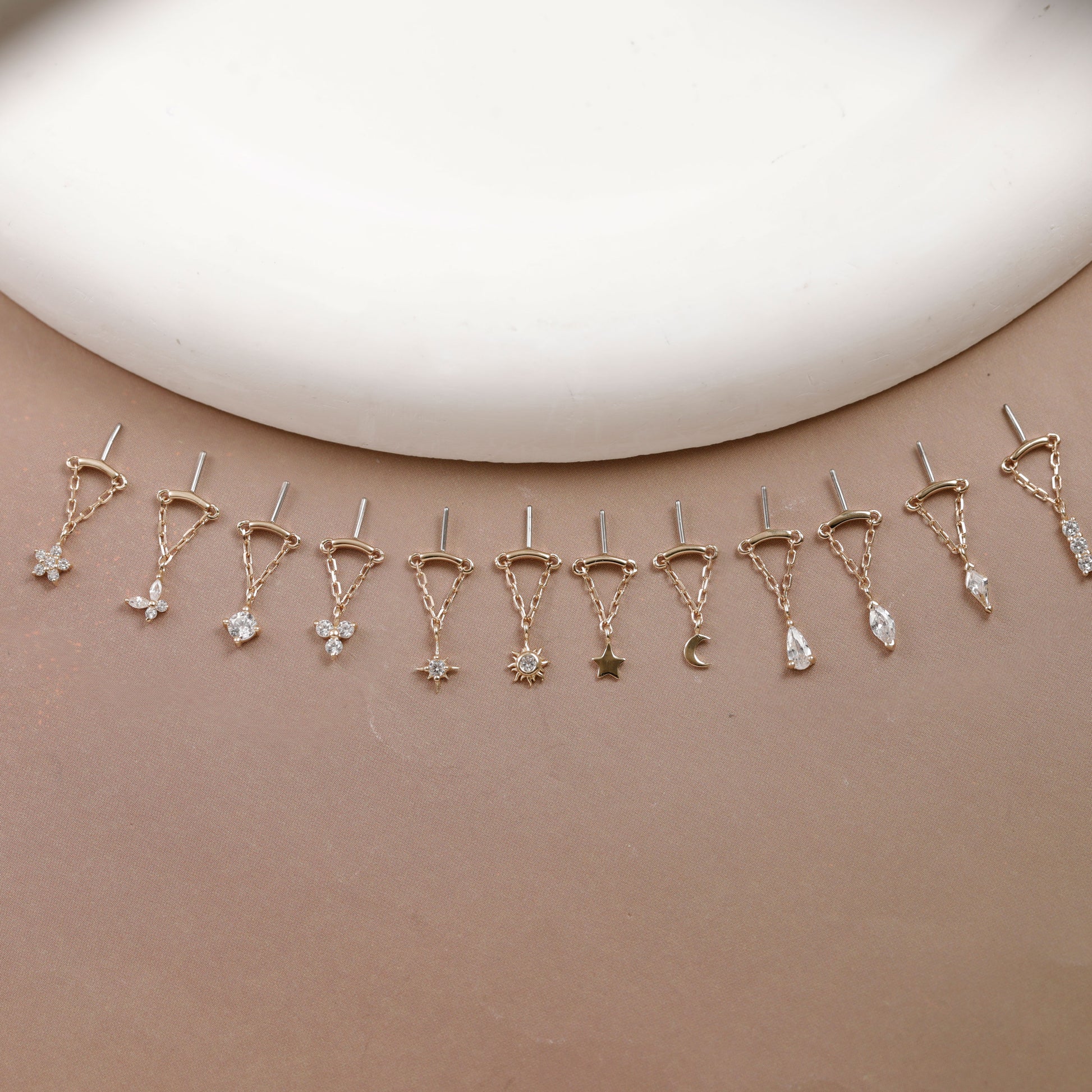 A set of gold-toned ear cuffs with dangling charms in various shapes including stars, moons, flowers, and teardrops, displayed on a neutral beige surface with a white curved prop at the top.