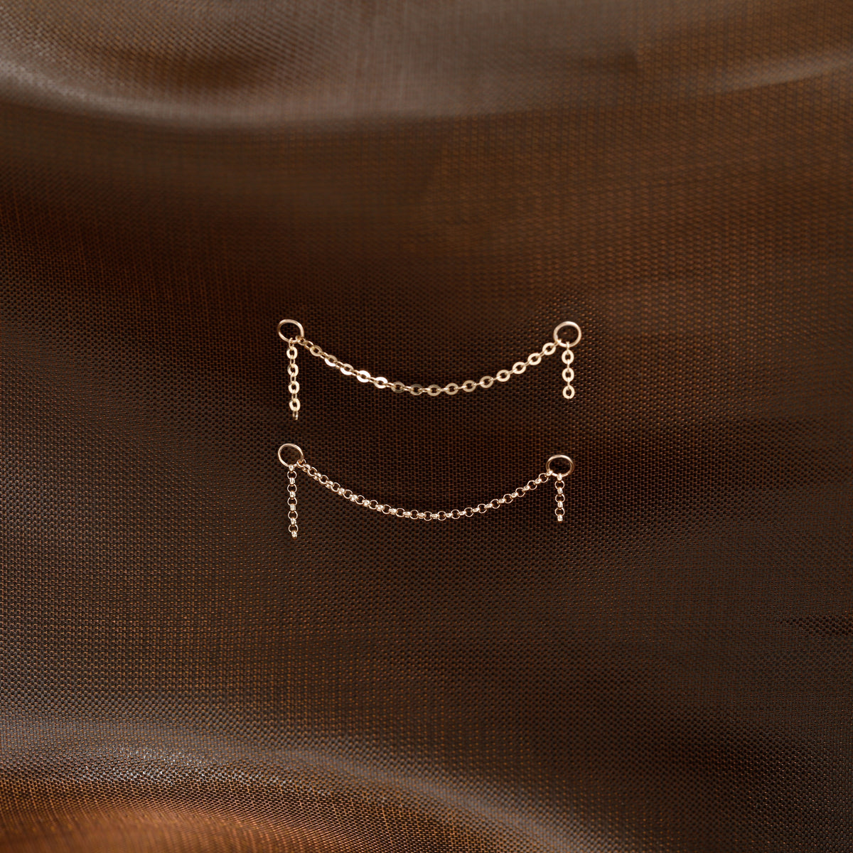 Two small gold chain link earrings with dangling loops, displayed on a textured dark brown fabric background.