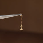A tiny gold dangling charm with two small fan-like shapes, held by metal tweezers against a dark brown background.