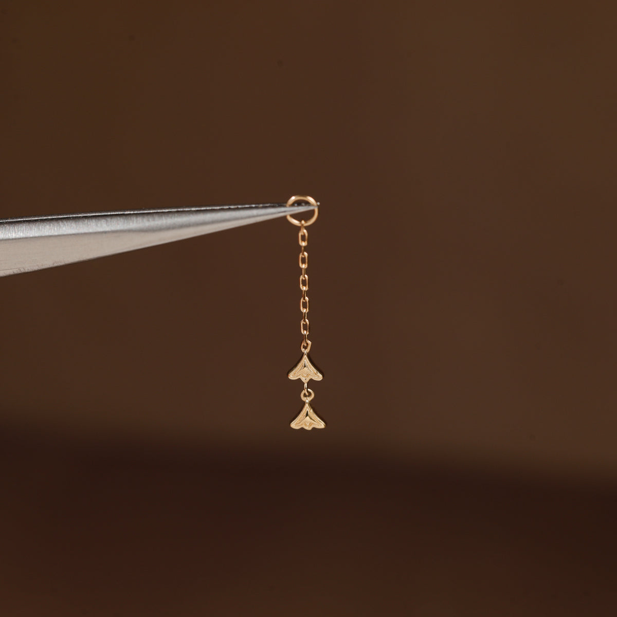 A tiny gold dangling charm with two small fan-like shapes, held by metal tweezers against a dark brown background.