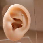 A delicate gold ear cuff with a small round stud and a short chain leading to a small triangular charm, displayed on a beige silicone ear model against a soft warm brown background.