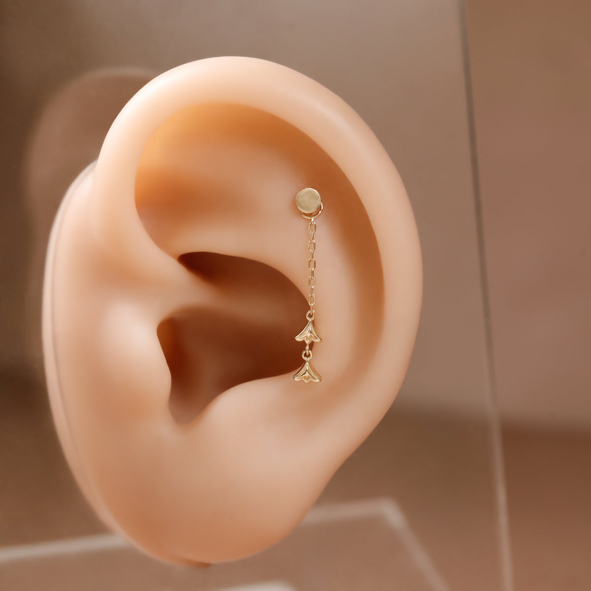 A delicate gold ear cuff with a small round stud and a short chain leading to a small triangular charm, displayed on a beige silicone ear model against a soft warm brown background.
