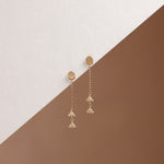 A pair of gold dangle earrings with circular studs, short chain links, and three dangling fan-shaped charms, displayed on a diagonally split white and brown background.