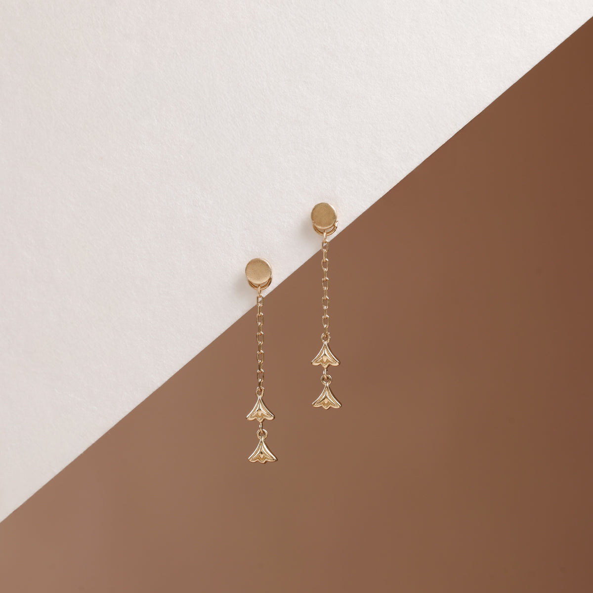 A pair of gold dangle earrings with circular studs, short chain links, and three dangling fan-shaped charms, displayed on a diagonally split white and brown background.