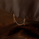 A pair of delicate rose gold hoop earrings with a short chain detail, shown hanging against a dark brown sheer fabric backdrop.