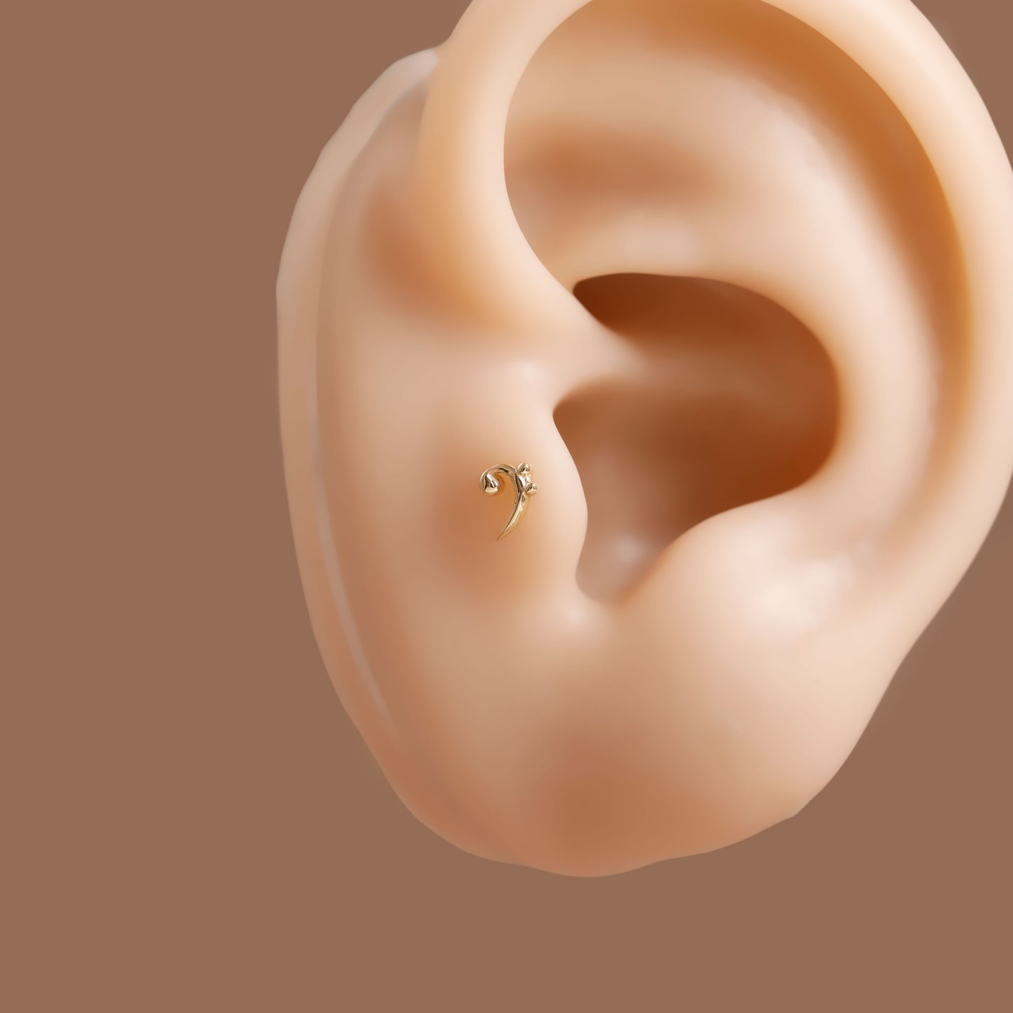 A small gold ear piercing stud shown on a realistic beige ear model against a solid brown background.