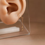 A delicate gold hoop earring with a short chain and two small triangular dangling charms, shown worn on a beige ear mannequin against a warm beige background.