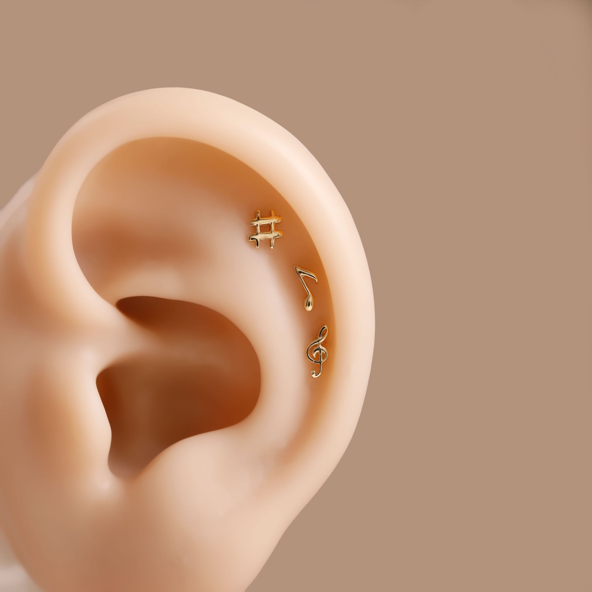 Three small gold ear piercings shaped like a hashtag, a music note, and a treble clef, shown on a beige ear model against a solid taupe background.