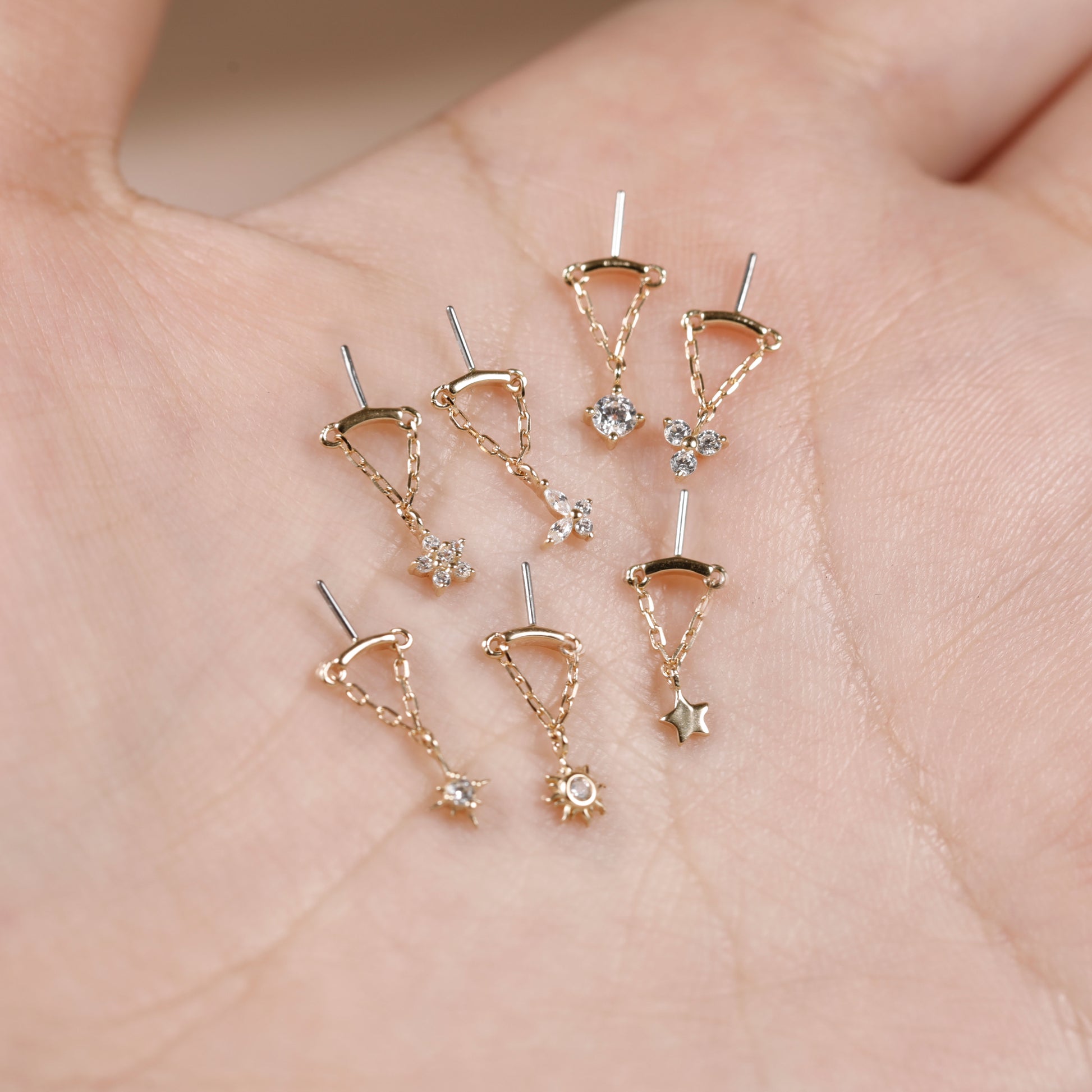A set of nine delicate gold-toned dangle earrings displayed on an open palm. The earrings feature various celestial and geometric charms, including stars, moons, suns, and triangles, some adorned with clear rhinestones.