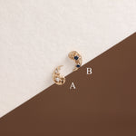 Two gold crescent moon stud earrings with blue and clear gemstones, labeled A and B on a split cream and brown background.