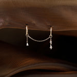 A pair of gold hoop earrings with a delicate chain connecting two small dangling charms, displayed on a brown textured fabric background.