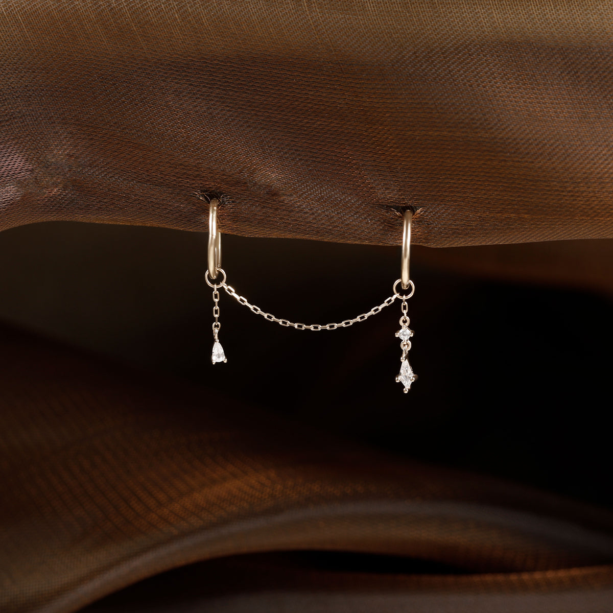 A pair of gold hoop earrings with a delicate chain connecting two small dangling charms, displayed on a brown textured fabric background.