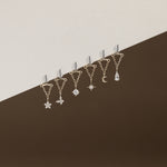 Six gold-toned ear cuffs with dangling celestial charms, including a star, flower, gem, moon, and teardrop, arranged diagonally against a split beige and dark brown background.