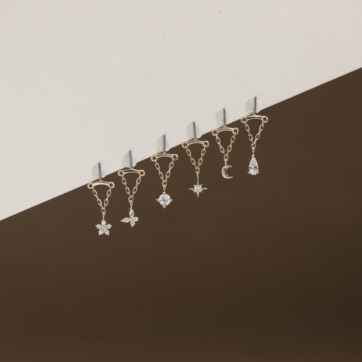 Six gold-toned ear cuffs with dangling celestial charms, including a star, flower, gem, moon, and teardrop, arranged diagonally against a split beige and dark brown background.