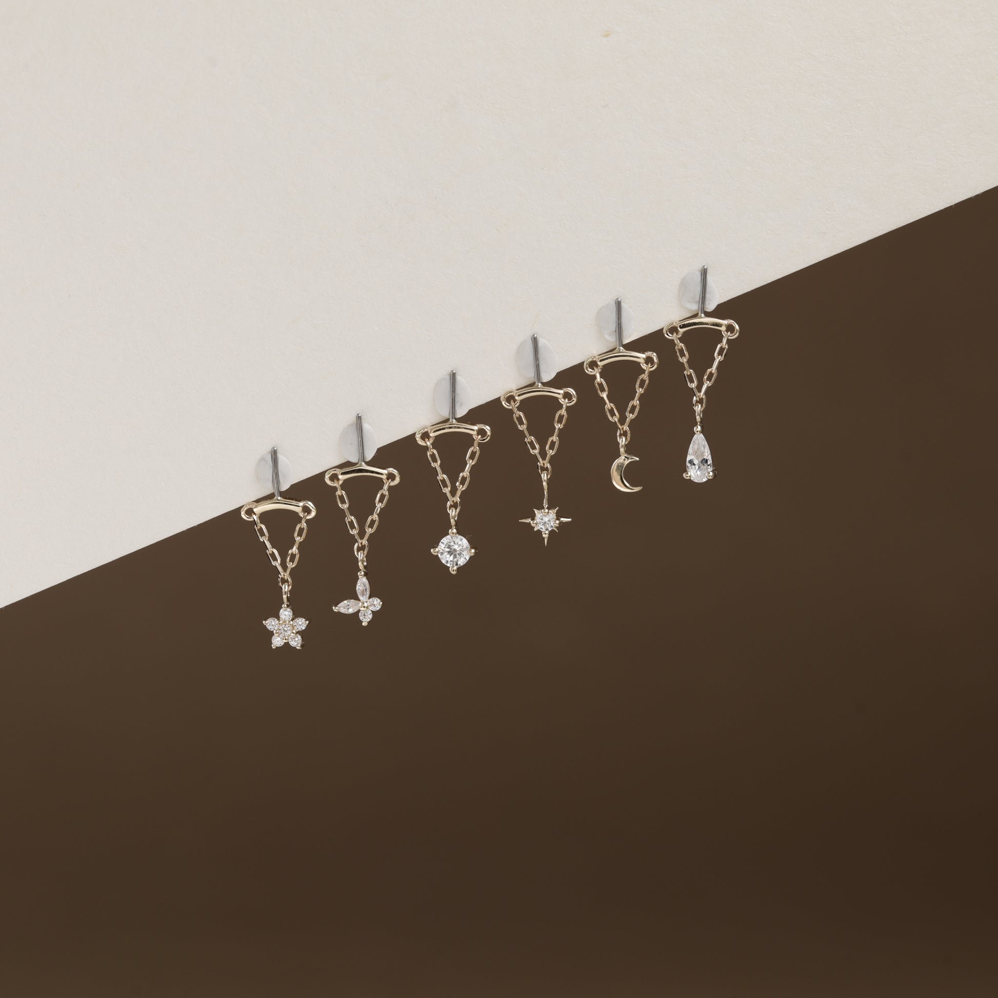 Six gold-toned ear cuffs with dangling celestial charms, including a star, flower, gem, moon, and teardrop, arranged diagonally against a split beige and dark brown background.