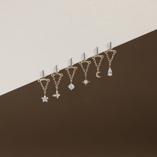Six gold-toned ear cuffs with dangling celestial charms, including a star, flower, gem, moon, and teardrop, arranged diagonally against a split beige and dark brown background.