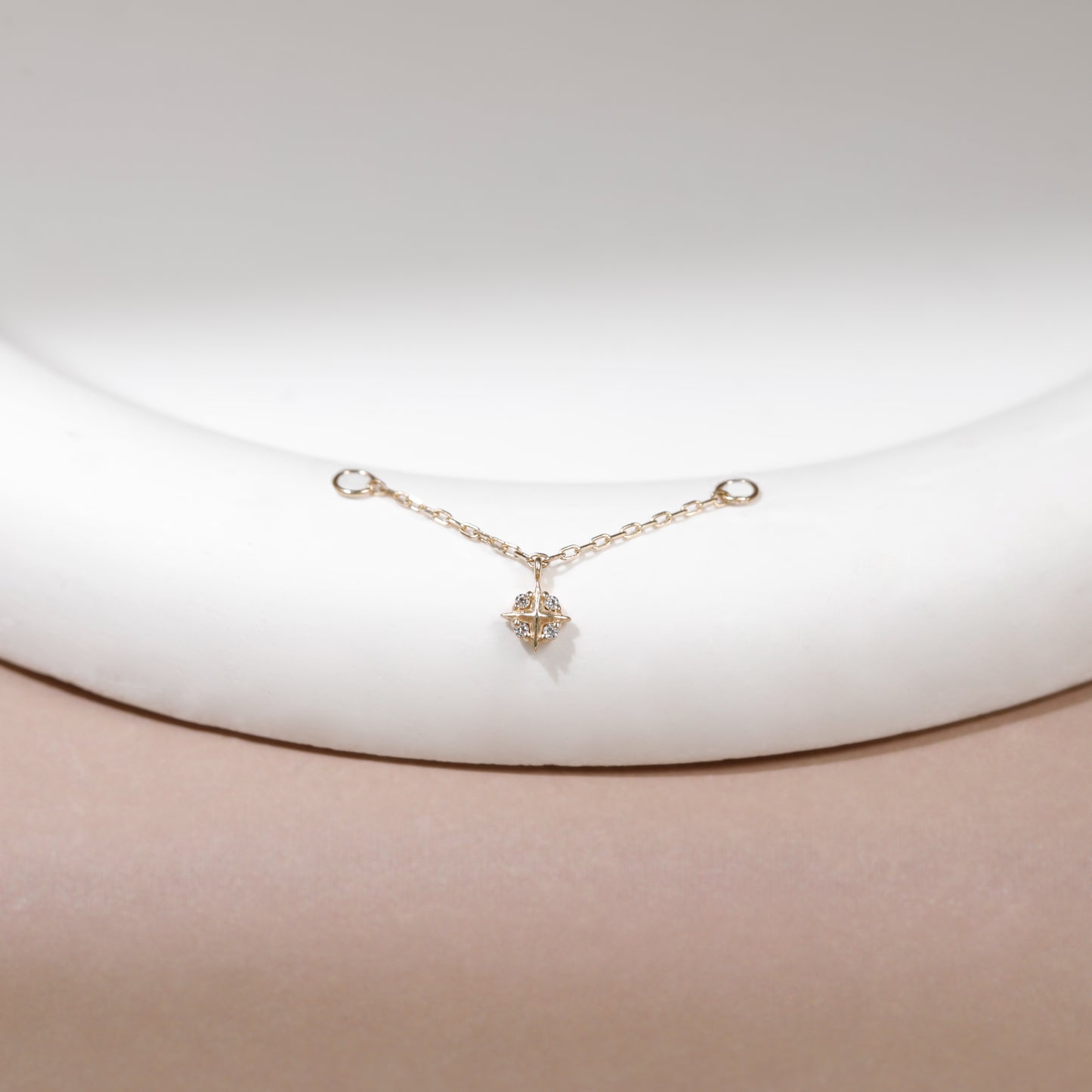 A delicate gold chain bracelet with a small geometric star-like charm, displayed on a white curved jewelry stand against a soft beige background.