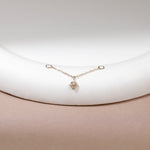 A delicate gold chain bracelet with a small geometric star-like charm, displayed on a white curved jewelry stand against a soft beige background.