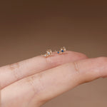 A pair of small gold crescent moon stud earrings with tiny clear stones and a single blue gemstone, shown resting on a person's fingers against a soft brown background.