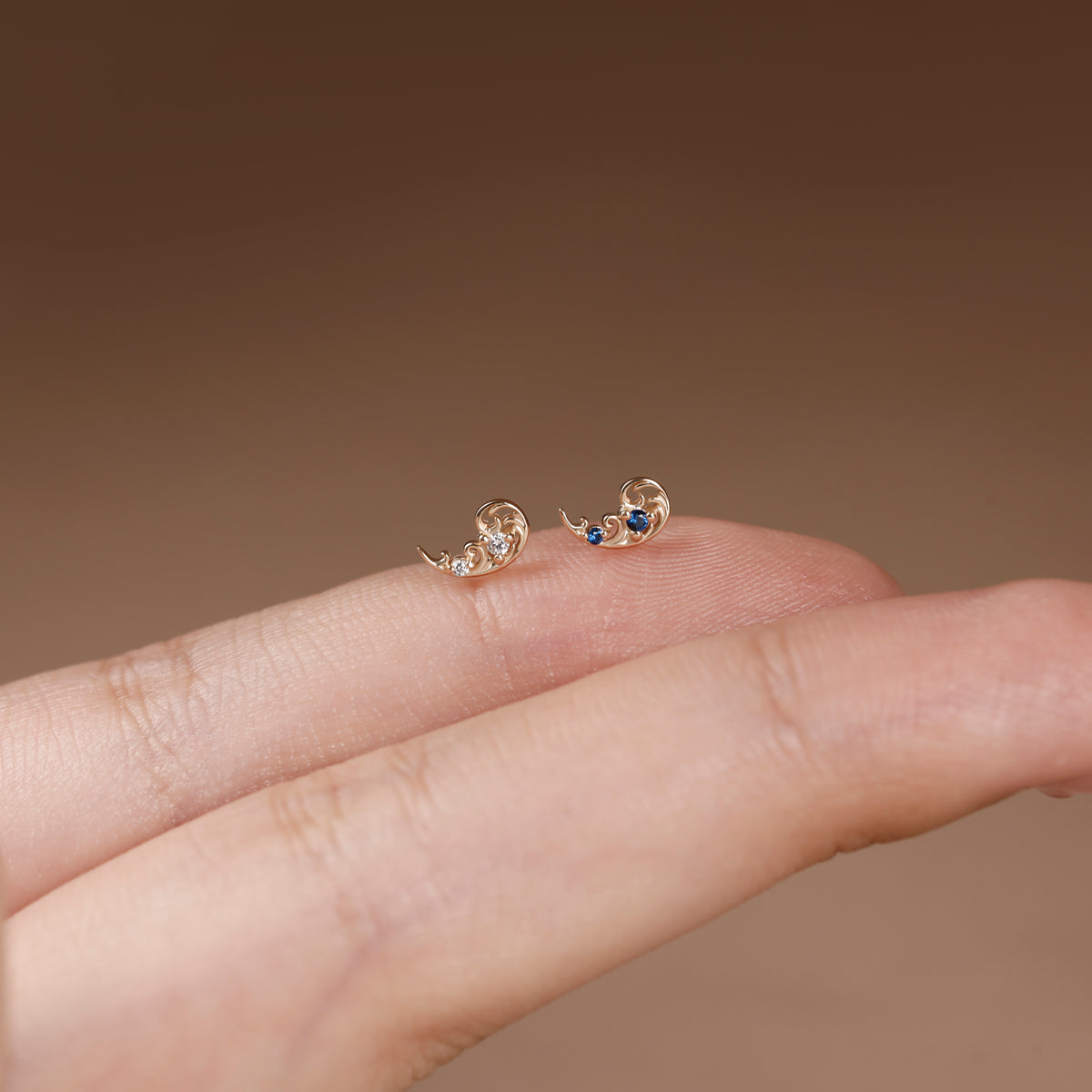 A pair of small gold crescent moon stud earrings with tiny clear stones and a single blue gemstone, shown resting on a person's fingers against a soft brown background.
