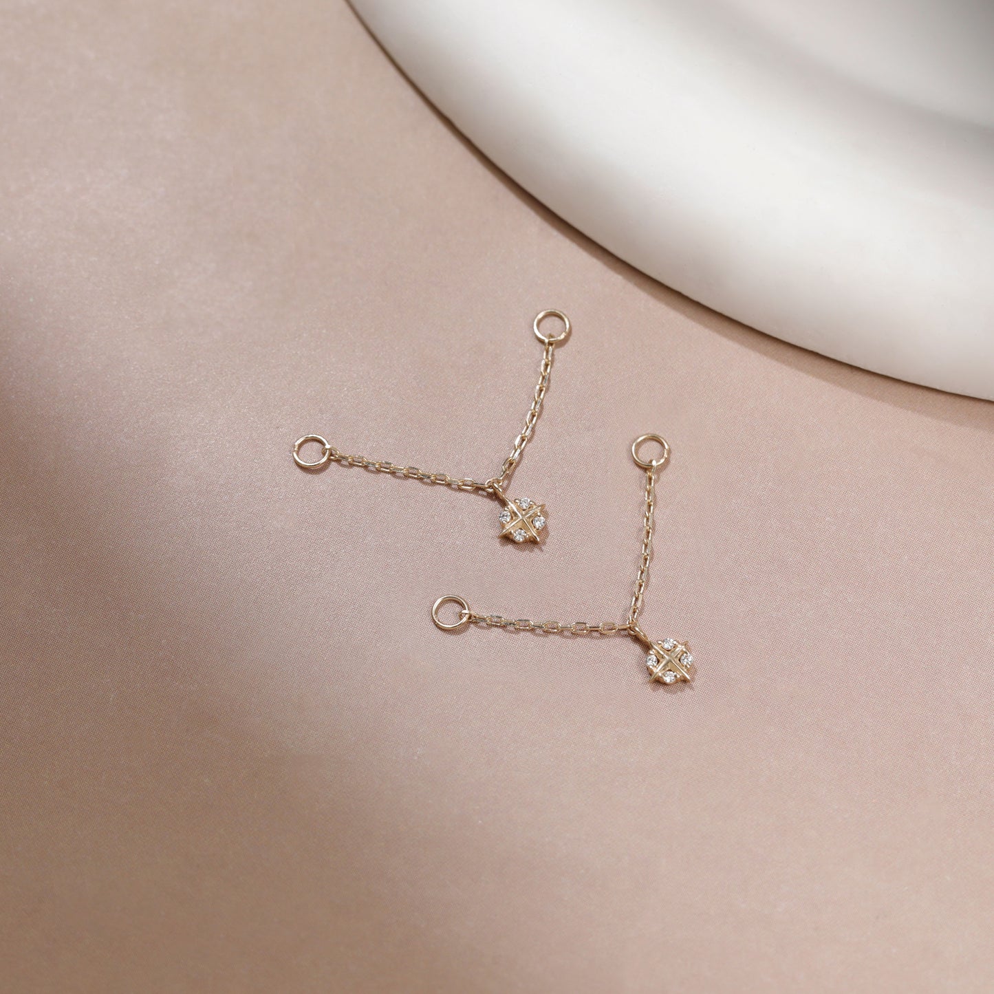 A pair of delicate gold chain earrings with small floral charms, displayed on a soft beige surface with a white curved prop in the background.