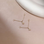 A pair of delicate gold chain earrings with small floral charms, displayed on a soft beige surface with a white curved prop in the background.