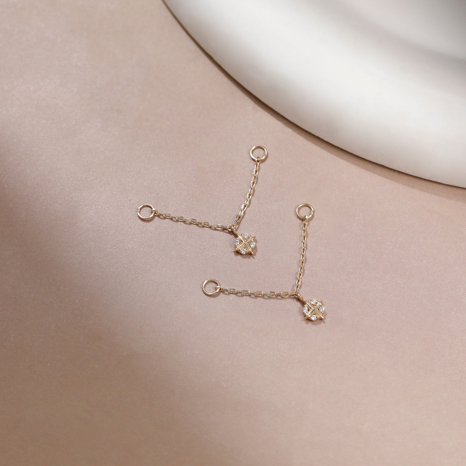 A pair of delicate gold chain earrings with small floral charms, displayed on a soft beige surface with a white curved prop in the background.