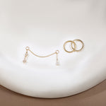 A pair of gold-toned earrings displayed on a white curved surface, including two interlocking hoop-style rings and a delicate chain bridge with small dangling clear crystal drops.